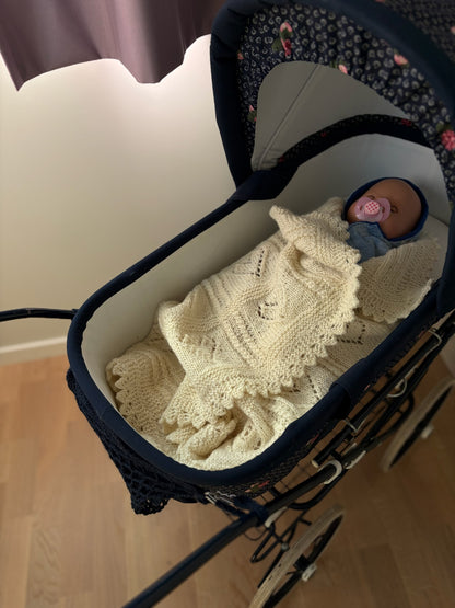 Baby ligger i en mørk barnevogn under et kremhvitt Hjertebank-babyteppe.