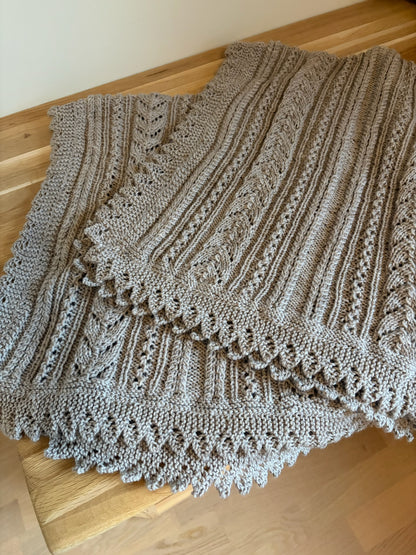 Lys beige Ingrid-babyteppe med hullmønster og dekorativ blondekant ligger på et trebord.