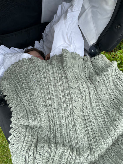 Baby ligger i en svart barnevogn under et grågrønt Ingrid-babyteppe.