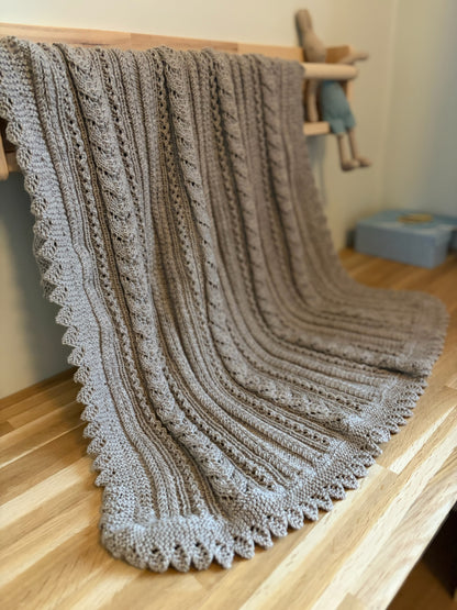 Lys beige Ingrid-babyteppe henger over et lyst trestativ med mønsteret synlig.