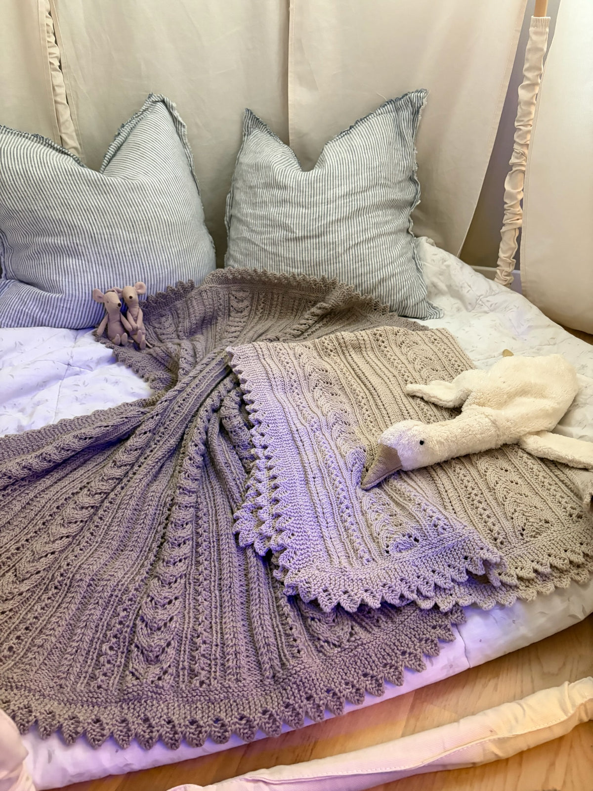 Lilla og beige Ingrid-babytepper ligger på en seng med puter og et hvitt kosedyr.