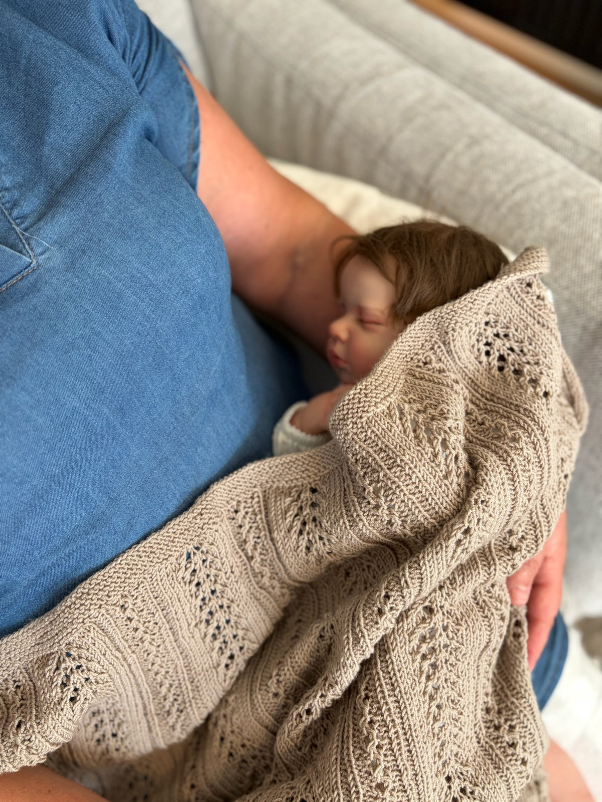 Sovende barn ligger på fanget til en voksen, pakket inn i et beigefarget Magnus-babyteppe.