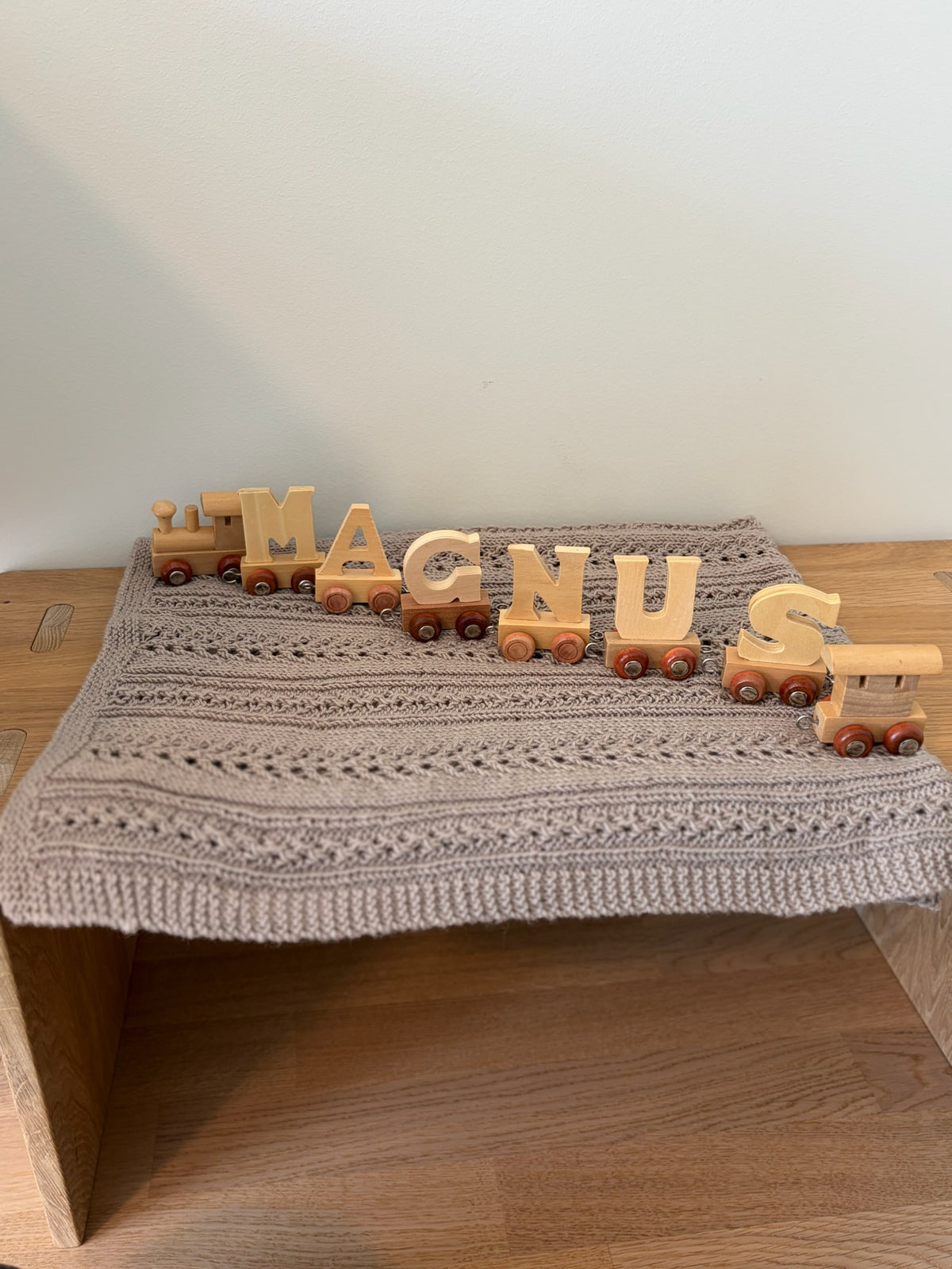 Beigefarget Magnus-babyteppe på en trebenk med et leketog som staver navnet Magnus.