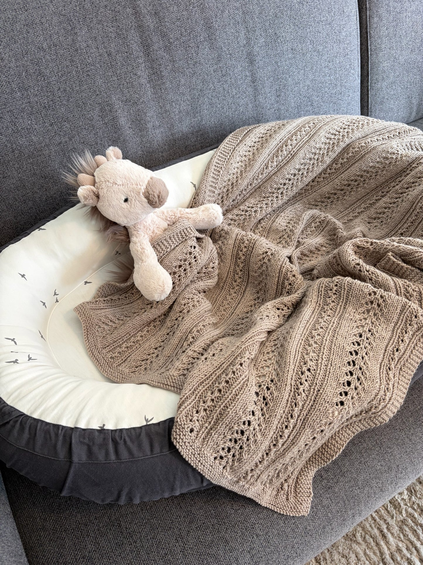 Beigefarget Magnus-babyteppe ligger over et babynest i en grå sofa sammen med et kosedyr.