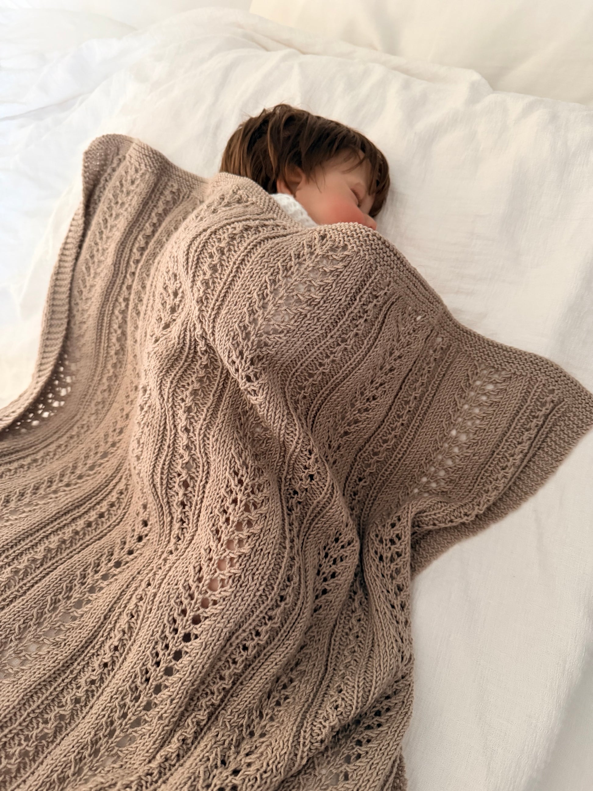 Sovende barn under et beigefarget Magnus-babyteppe i en hvit seng.