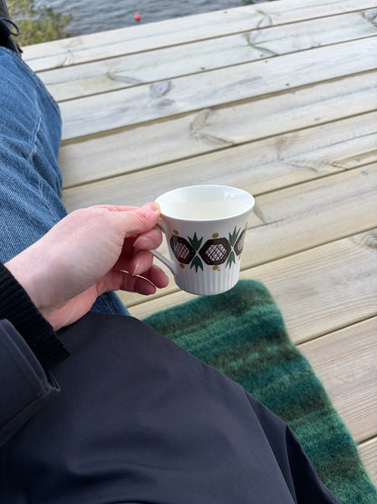Person sitter på et grønt Ton i Ton-sitteunderlag og holder en kopp på en treterrasse.